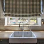 A kitchen sink area with a white double basin, light wood countertops, and a plaid Roman shade over a large window looking out to a backyard pool.