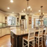A traditional kitchen with cream-colored cabinetry, a large granite island with white wooden barstool seating, and elegant hanging chandeliers.
