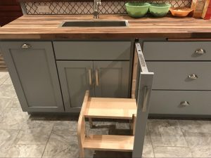 A close-up of grey kitchen cabinetry featuring a pull-out wooden step stool integrated into a slim vertical drawer next to the sink.