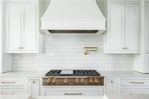 A high-end white kitchen featuring a professional gas range, a white custom vent hood, and a gold wall-mounted pot filler. Image Description: This chef-inspired setup combines style and function with a professional-grade range and a pot filler, allowing you to fill heavy pots directly on the stove.