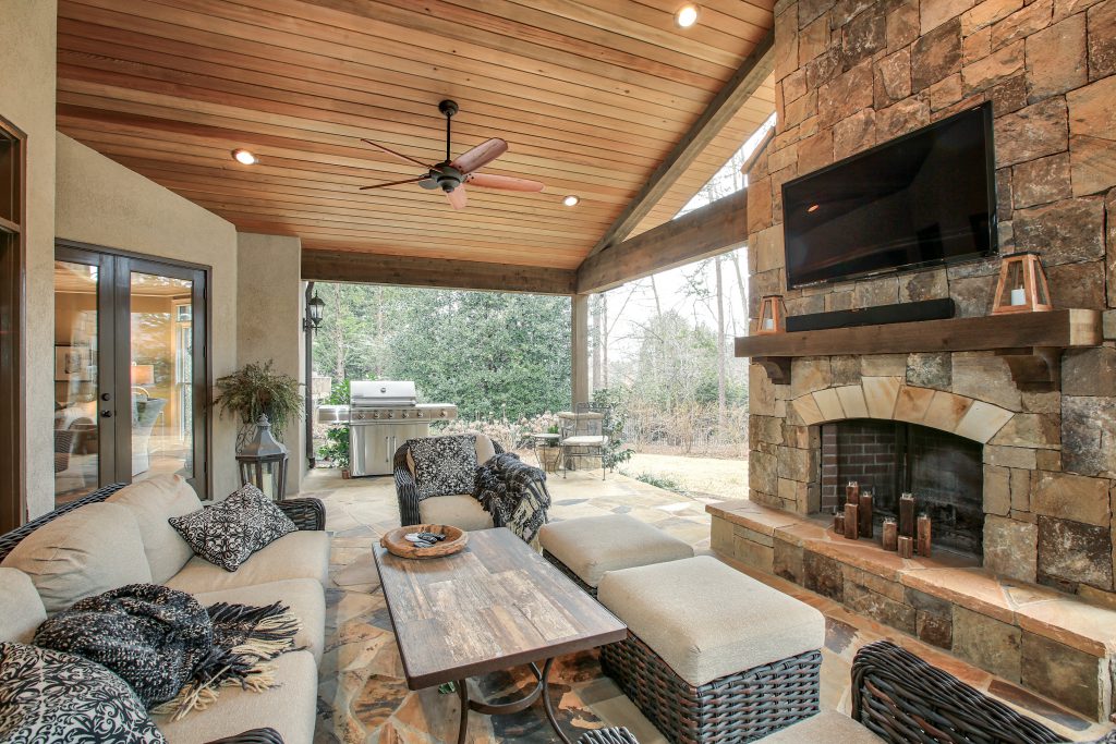 High-end outdoor living pavilion with a massive stone fireplace, mounted TV, outdoor kitchen, and comfortable patio seating.

